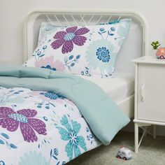 a white bed with blue and purple flowers on the comforter next to a night stand