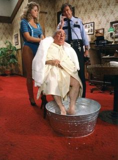 an old man getting his hair brushed by two women in a room with red carpet