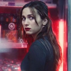 a woman with long hair standing in front of a red neon light and looking at the camera