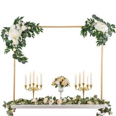 an arrangement of flowers, candles and greenery on a white table with a gold frame