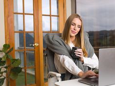 a woman sitting at a desk with her laptop and holding a cup in front of her