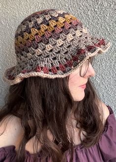 a woman with long hair wearing a crocheted hat and purple top, standing against a wall