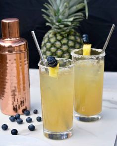 two glasses filled with lemonade and blueberries sit on a table next to a pineapple