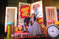 a man and woman sitting on top of a giant clock in front of colorful posters