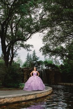 a woman in a purple dress is standing near a pond with trees and water behind her