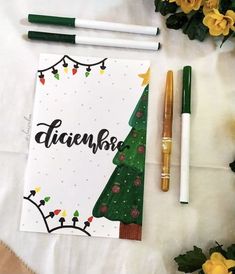 a notepad with christmas tree on it next to two pens and some yellow flowers