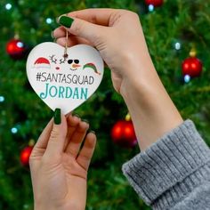 someone holding up a heart shaped ornament in front of a christmas tree that says, santasquad jordan