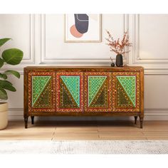 an ornately decorated sideboard with green and red designs on the doors, in front of a potted plant