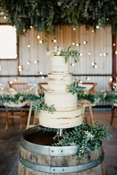 a wedding cake sitting on top of a wooden barrel in front of tables with greenery