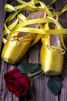 a pair of yellow ballet shoes next to a red rose on a wooden surface with ribbon