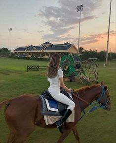 a woman riding on the back of a brown horse across a lush green field at sunset