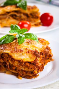 two white plates with lasagna on them and tomatoes, basil leaves, and cherry tomatoes in the background