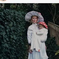 a woman holding a baby wearing a white dress and red polka dot hat while standing next to a hedge