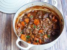 a pot filled with beef and carrots on top of a wooden table next to a white casserole dish