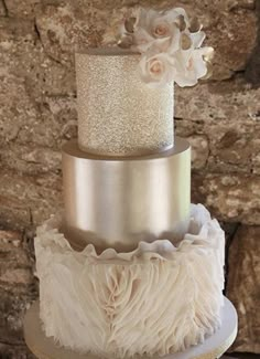 a three tiered cake with white flowers and feathers on top, sitting in front of a stone wall