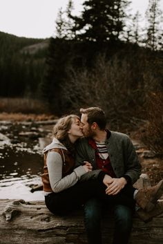 a couple kissing while sitting on a log in front of a river and forest during the day