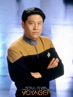 a man with his arms crossed standing in front of a blue and yellow background wearing a star trek uniform