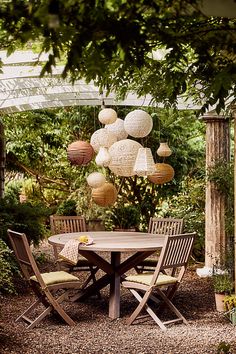 a wooden table surrounded by chairs and hanging paper lanterns in a garden with gravel ground