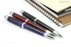 three pens sitting next to each other on top of a book and notebook with spiral bound pages