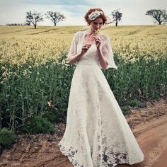 a woman in a white dress standing on a dirt road near a field with yellow flowers