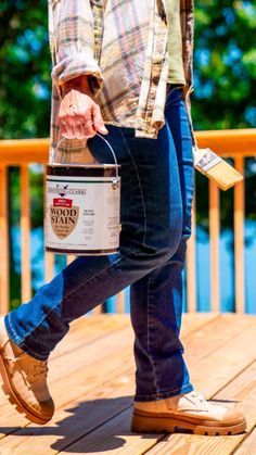 a person holding a paint bucket and walking on a wooden deck with trees in the background