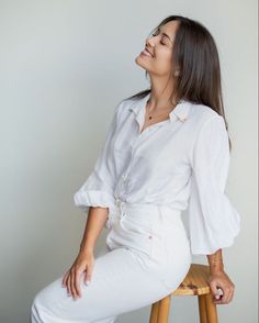 a woman sitting on top of a wooden stool next to a white wall and smiling