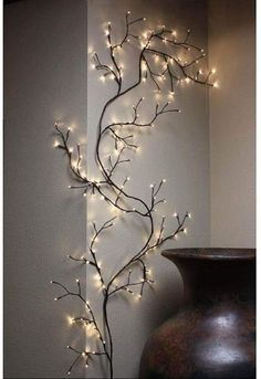 a vase sitting next to a white wall with lights on it and a tree branch in the corner