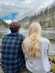 a man and woman sitting on rocks looking out over a river with mountains in the background