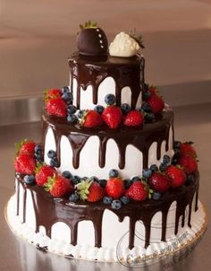 a triple layer cake with chocolate icing, strawberries and blueberries on top