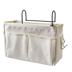 a white canvas storage bag with two handles and three pockets on the front, hanging from a black metal hook