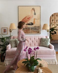 a woman walking through a living room with pink furniture and paintings on the wall behind her