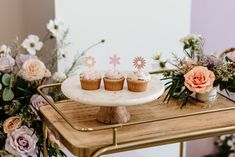 three cupcakes sitting on top of a cake stand with flowers in the background