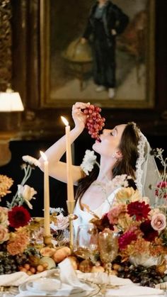 a woman in a wedding dress is eating grapes at a table with flowers and candles