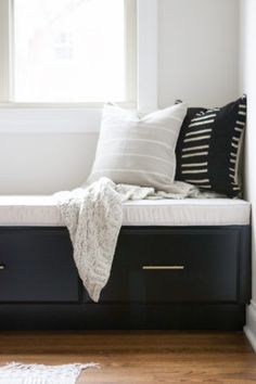 a window seat with pillows on it in front of a white wall and wooden floor