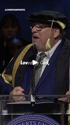 a man standing at a podium in front of a microphone wearing a hat and gown