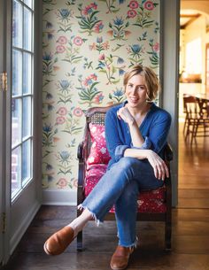 a woman sitting on a chair in front of a wallpapered room with flowers