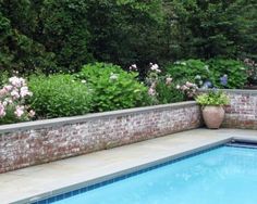 a swimming pool surrounded by flowers and greenery next to a brick wall with a planter in the middle