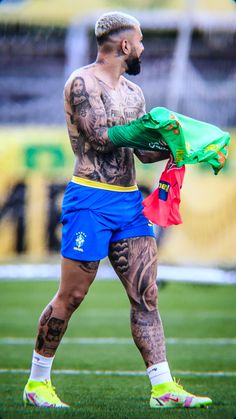 a tattooed man holding a green jacket on top of a soccer field with other players in the background