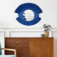 an eye shaped mirror hangs on the wall above a blue chair and wooden dresser in a white room