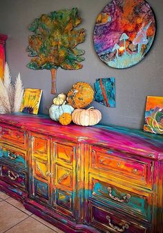 a colorful dresser with paintings on the wall behind it and two trees in the background