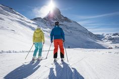 two people on skis are standing in the snow near a large mountain with a bright sun behind them