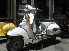 a white scooter is parked in front of a building with two yellow mopeds