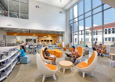 people are sitting in the library with orange and white chairs, tables, and bookshelves