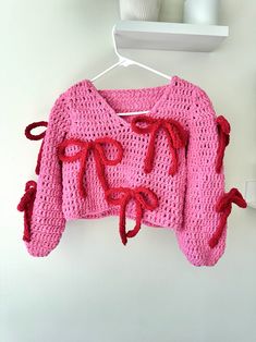 a pink sweater with red bows hanging on a white hanger next to a cup