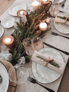 the table is set with white plates and silverware, pine cones, greenery, candles and napkins