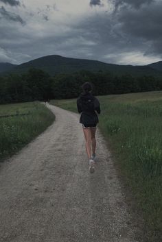 a woman running down a dirt road in the middle of a field with mountains in the background