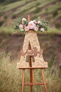 a wooden easel with flowers and writing on it in front of a grassy hill