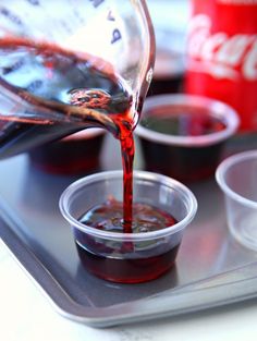 someone pouring red liquid into small cups on a tray with coca - cola cans in the background