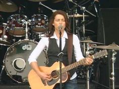 a woman singing into a microphone while holding an acoustic guitar in front of a drum set