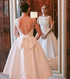 a woman standing in front of a mirror wearing a white dress with a bow on the back